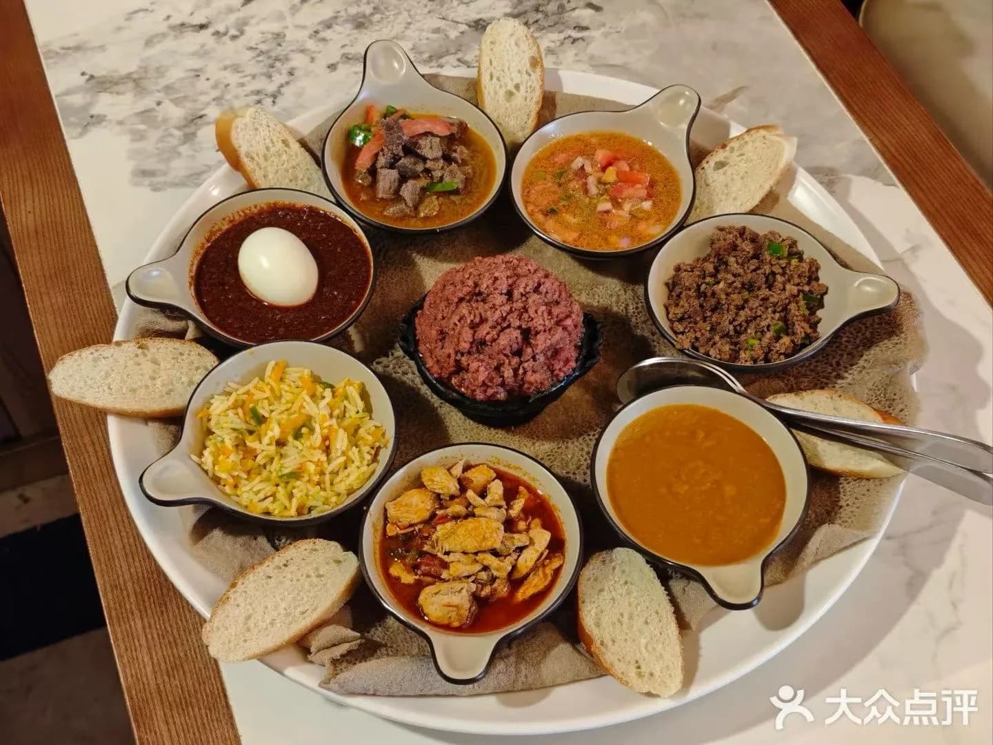 A round shared platter with several small dishes arranged over injera and slices of bread around the edge.