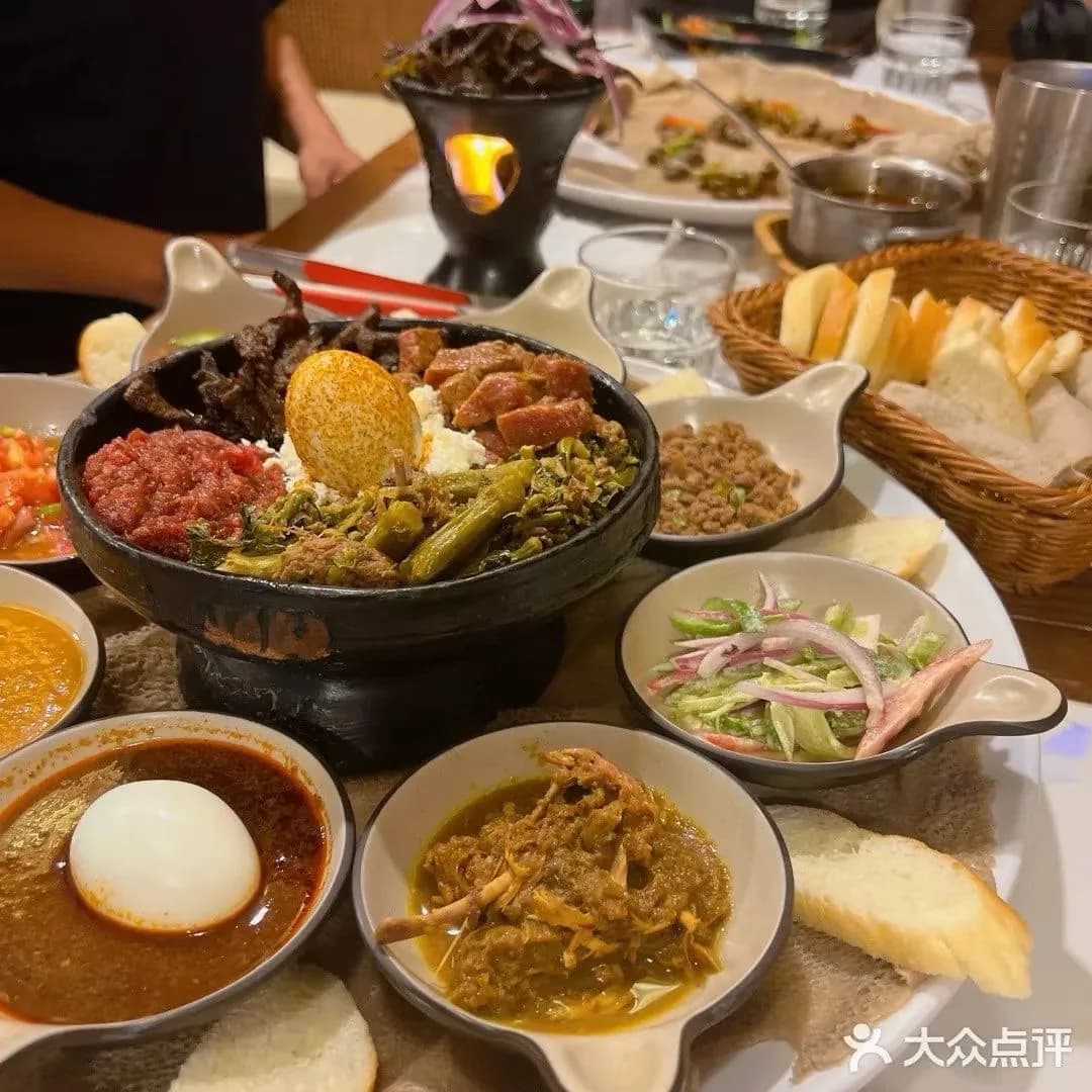 A shared restaurant table with a central black serving bowl, side dishes, injera, and bread.