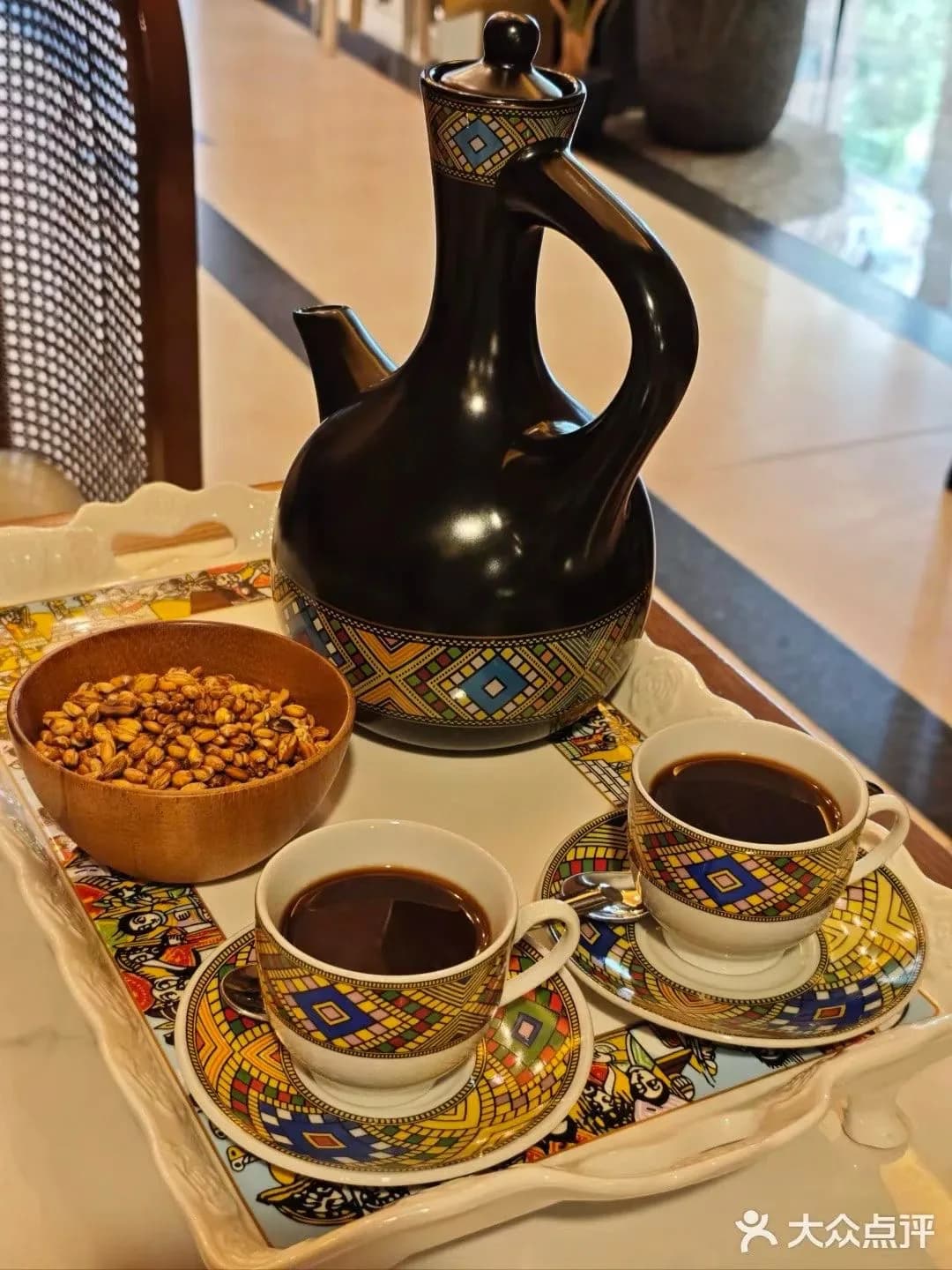 A decorated black Ethiopian coffee pot, two patterned cups of coffee, and a wooden bowl of snacks on a tray.