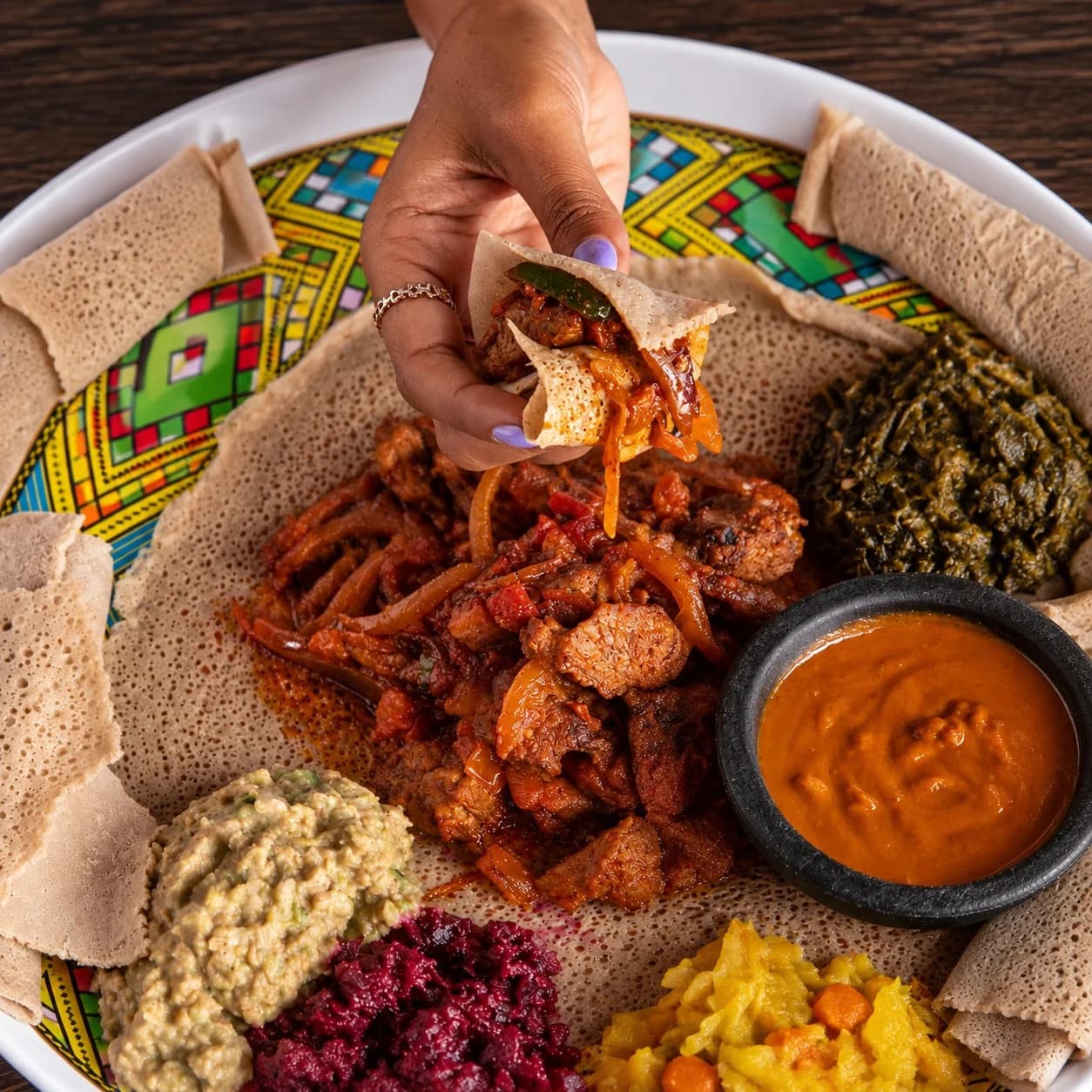 A hand holding a folded piece of injera filled with stew above a shared platter.